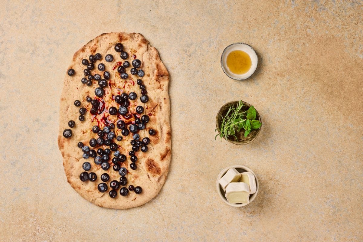 Close-up of a blueberry flatbread topped with fresh berries, surrounded by honey, herbs, and butter on a textured surface.