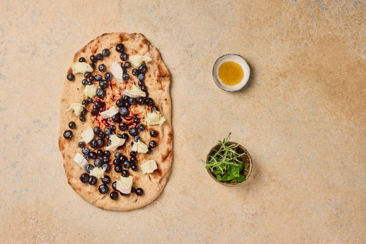 A flatbread blueberry pizza topped with blueberries, cheese, and herbs on a textured surface.