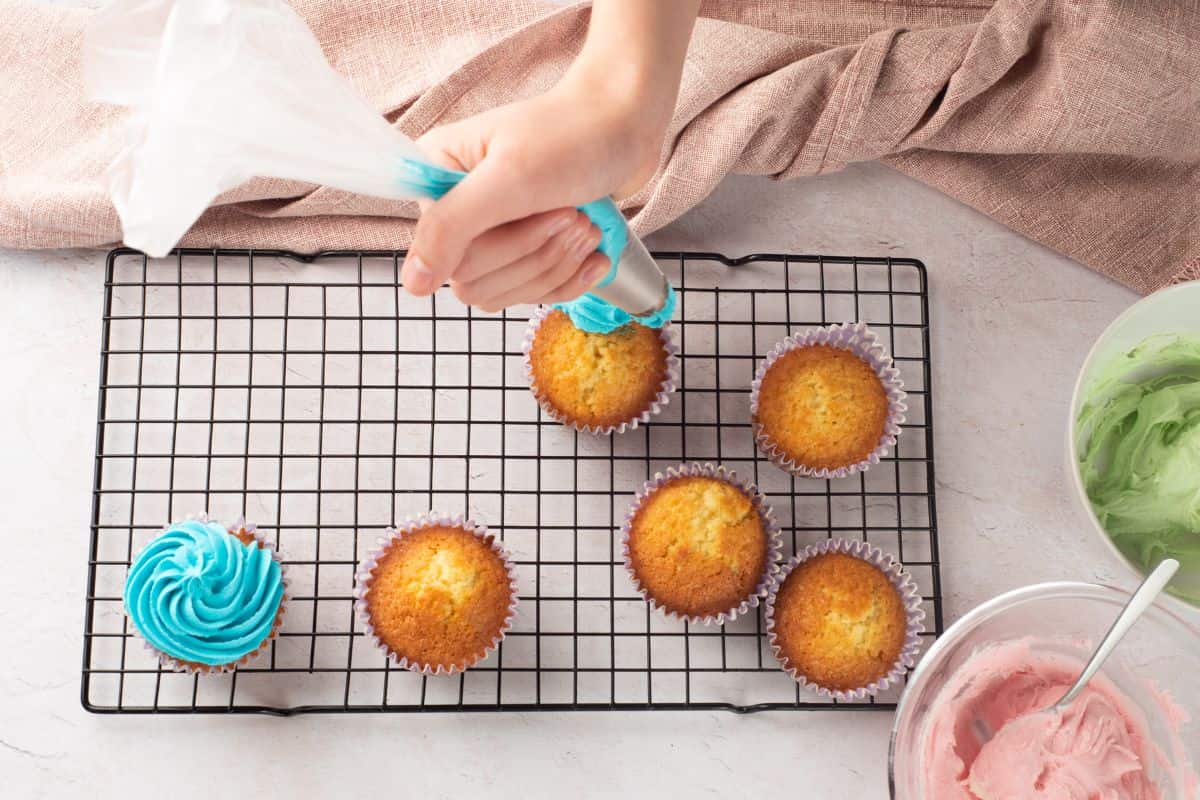 A hand decorates vanilla cupcakes with blue icing on a wire rack, surrounded by pastel frosting bowls.